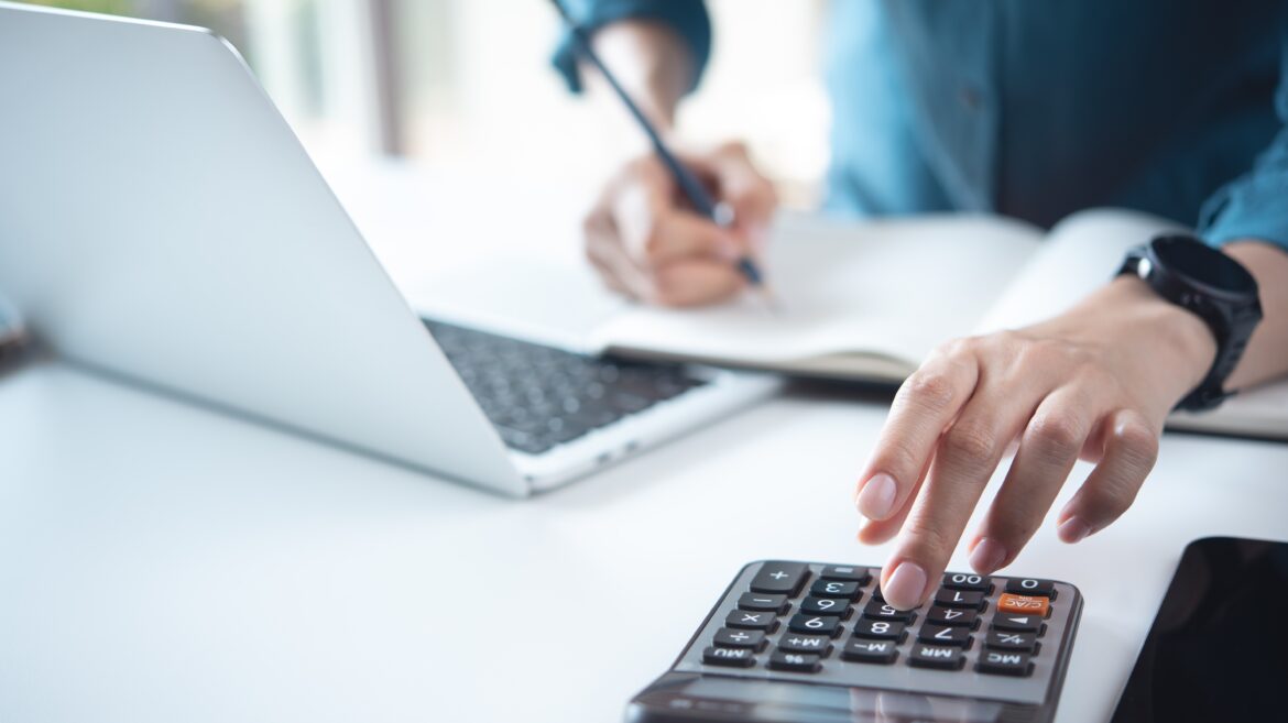 Person calculating taxes using a calculator, laptop and notepad, reviewing income, ISAs and pension contributions to plan tax-efficient investing.
