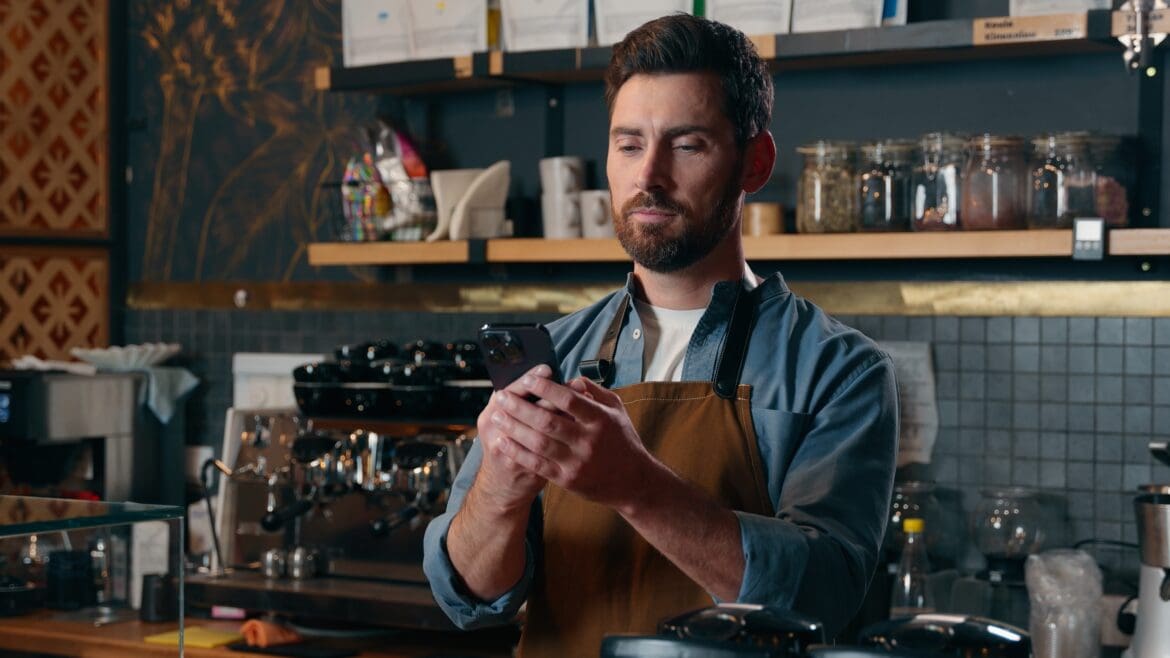 A small business owner in an apron checks his phone at the counter of his café, considering how to invest his business cash.